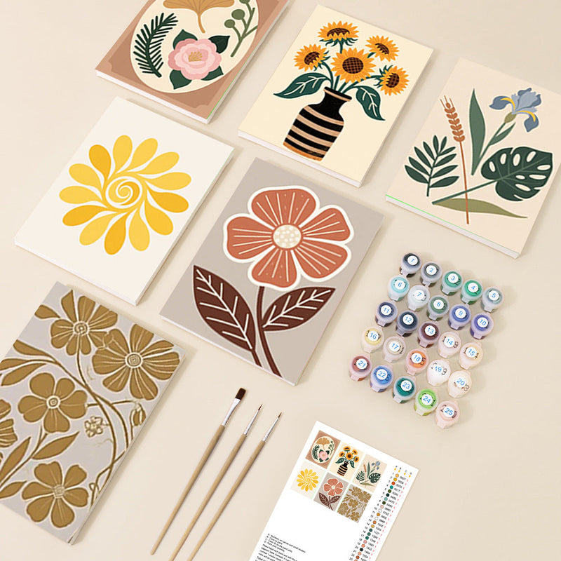 An overhead view of multiple small canvas boards displaying various modern boho floral and botanical designs, along with numbered paint pots and brushes, ready for a paint-by-number activity.